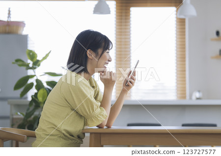 一位 30 多歲的女人微笑著坐在家中的餐桌旁看著自己的智慧型手機螢幕 123727577
