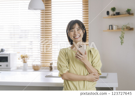 一位 30 多歲的女性在自家廚房裡微笑著用智慧型手機看著鏡頭 123727645