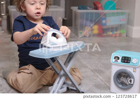 Little boy plays with toy iron and washing machine at home on floor. Cleaning, household chores, kids and educational games. Little boy plays with toy iron and washing machine at home on floor. Cleaning, household chores, kids and educational games. 123728617