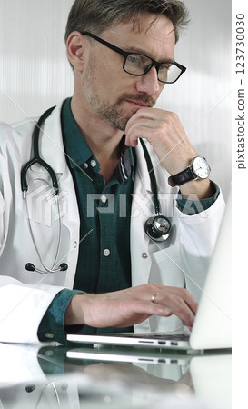 A male doctor in a green shirt under a medical coat is thinking while using technology in healthcare. Man is typing on a laptop on a glass desk in the clinic, vertical portrait view. Medicine concept 123730030