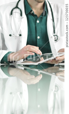 Doctor man in green shirt under a white coat using digital tablet in modern healthcare setting, interacting with patient data. Medicine concept Doctor man in green shirt under a white coat using digital tablet in modern healthcare setting, interacting with patient data. Medicine concept 123730035