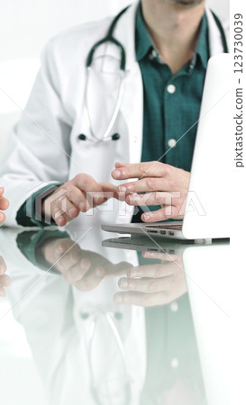 Doctor man wearing a stethoscope and lab coat is gesturing with hands while explaining a diagnosis to a patient across a desk with a laptop. Medicine concept Doctor man wearing a stethoscope and lab coat is gesturing with hands while explaining a diagnosis to a patient across a desk with a laptop. Medicine concept 123730039