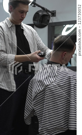 Professional barber grooming client's hair using electric clipper, delivering precise cut with skill in contemporary salon setting Professional barber grooming client's hair using electric clipper, delivering precise cut with skill in contemporary salon setting 123730052