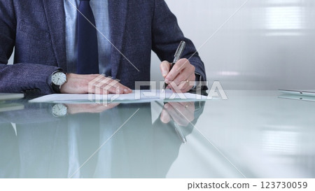 Businessman in a professional attire signing a contract with a pen on a modern glass desk in a contemporary office environment. Business people or audit concept Businessman in a professional attire signing a contract with a pen on a modern glass desk in a contemporary office environment. Business people or audit concept 123730059