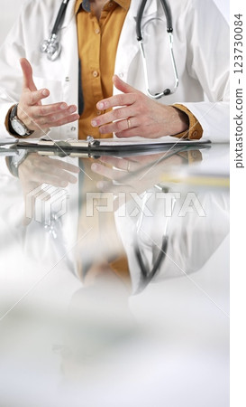 Unknown male doctor wearing a stethoscope, yellow shirt under a lab coat is gesturing with hands overs a glass desk with a laptop in clinic office. Medicine concept 123730084