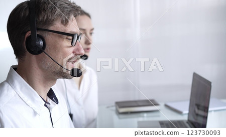 Happy smiling male call center operator in white casual shirt is assisting clients online, providing customer support and answering inquiries. Business call center operators Happy smiling male call center operator in white casual shirt is assisting clients online, providing customer support and answering inquiries. Business call center operators 123730093