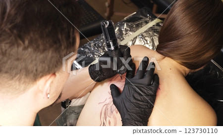 Professional tattoo artist wearing black gloves drawing intricate design on woman's back while working in sleek, well lit tattoo studio with modern equipment 123730110