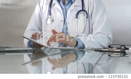 Doctor woman, wearing a blue blouse under a medical coat, is using digital tablet in modern healthcare setting, interacting with patient data, close-up. Medicine concept 123730132