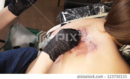 Professional tattoo artist wearing black gloves drawing intricate design on woman's back while working in sleek, well lit tattoo studio with modern equipment 123730133