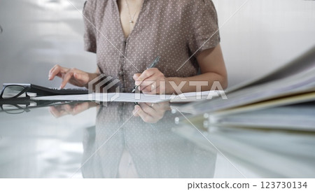 Businesswoman accountant, wearing brawn dotted blouse, is using a calculator to crunch numbers and jotting down meticulous notes, at sleek glass desk in a contemporary office. Taxes, audit in business 123730134