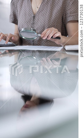 Businesswoman accountant in brawn dotted blouse reviewing financial reports with magnifying glass and calculator in office, vertical view. Taxes, audit in business Businesswoman accountant in brawn dotted blouse reviewing financial reports with magnifying glass and calculator in office, vertical view. Taxes, audit in business 123730138