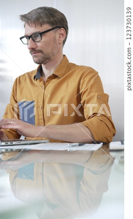 Focused businessman in yellow casual shirt is using laptop and thinking with hand on chin, working on a project in a modern office, vertical portrait. Business people concept 123730139