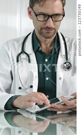 Doctor man in green shirt under a white coat using digital tablet in modern healthcare setting, interacting with patient data, vertical portrait. Medicine concept 123730149