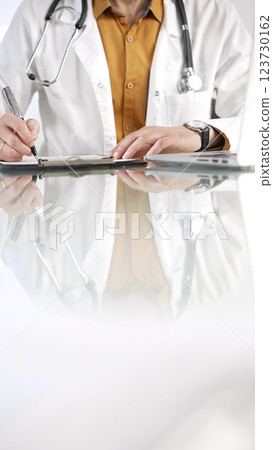 Unknown male doctor wearing a stethoscope, yellow shirt under a lab coat is using a clipboard and taking notes on glass desk in a medical office, close-up vertical view. Medicine concept Unknown male doctor wearing a stethoscope, yellow shirt under a lab coat is using a clipboard and taking notes on glass desk in a medical office, close-up vertical view. Medicine concept 123730162