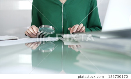 Businesswoman in green blouse carefully reviewing financial reports with magnifying glass and calculator, conducting audit and analyzing data in office. Taxes, audit in business 123730170