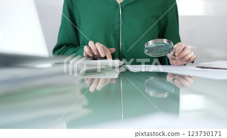 Businesswoman in green blouse carefully reviewing financial reports with magnifying glass and calculator, conducting audit and analyzing data in office. Taxes, audit in business Businesswoman in green blouse carefully reviewing financial reports with magnifying glass and calculator, conducting audit and analyzing data in office. Taxes, audit in business 123730171