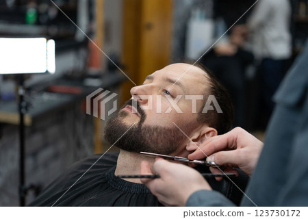 Skilled barber grooming clients beard using professional scissors, delivering precise styling within contemporary barbershop setting 123730172
