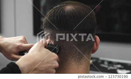 Skilled barber trimming male client's hair using electric clipper, creating contemporary hairstyle in modern urban barbershop setting with precise grooming technique, closeup 123730190