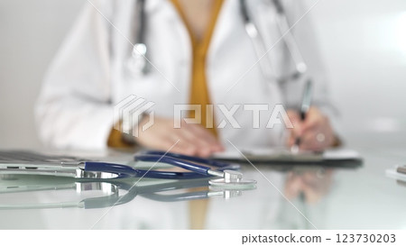 Stethoscope and medication history records form are on a glass table before a female doctor in yellow blouse under a white medical coat. Physician is working on clinical office background. Medicine 123730203
