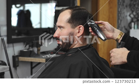 Skilled barber trimming male client's hair using electric clipper, creating contemporary hairstyle in modern urban barbershop setting with precise grooming technique 123730213