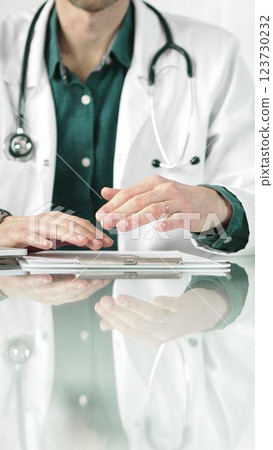 Doctor man wearing a stethoscope and lab coat is gesturing with hands while explaining a diagnosis to a patient across a glass desk with a laptop. Medicine concept 123730232