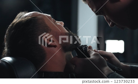 Skilled barber grooming client's beard using electric razor, delivering precise styling within contemporary barbershop setting 123730233