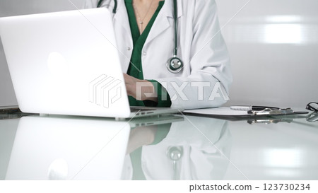 Female doctor using technology in healthcare, close up of hands typing on laptop with stethoscope, glasses, and clipboard on desk Female doctor using technology in healthcare, close up of hands typing on laptop with stethoscope, glasses, and clipboard on desk 123730234