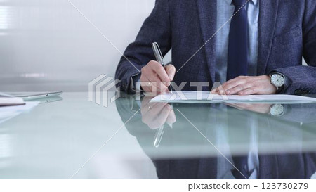 Businessman in a professional attire signing a contract with a pen on a modern glass desk in a contemporary office environment. Business people or audit concept Businessman in a professional attire signing a contract with a pen on a modern glass desk in a contemporary office environment. Business people or audit concept 123730279