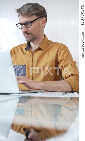 Focused businessman in yellow casual shirt is using laptop and thinking with hand on chin, working on a project in a modern office, vertical portrait. Business people concept 123730280
