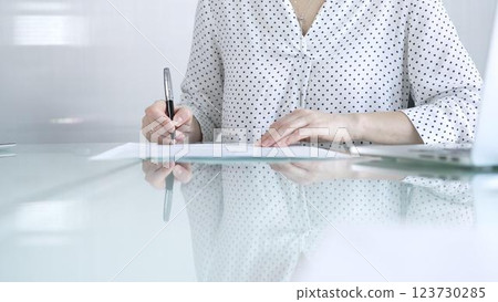 Businesswoman in white dotted blouse is signing a contract at a sleek glass desk in a modern office, showcasing professionalism and focus in her work environment. Business people concept 123730285