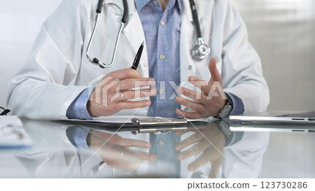 Male doctor wearing a blue shirt, stethoscope and lab coat is gesturing with hands while explaining a diagnosis to a patient across a glass desk with a laptop. Medicine concept Male doctor wearing a blue shirt, stethoscope and lab coat is gesturing with hands while explaining a diagnosis to a patient across a glass desk with a laptop. Medicine concept 123730286