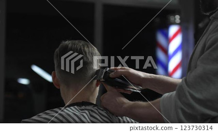 Skilled barber trimming client's hair with electric razor, creating precise contemporary hairstyle inside sleek urban barbershop setting Skilled barber trimming client's hair with electric razor, creating precise contemporary hairstyle inside sleek urban barbershop setting 123730314