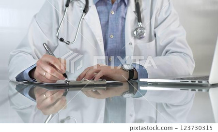 Male doctor, wearing blue shirt under a white medical coat, is using a clipboard and taking notes on glass desk in a medical office, close-up. Medicine concept Male doctor, wearing blue shirt under a white medical coat, is using a clipboard and taking notes on glass desk in a medical office, close-up. Medicine concept 123730315