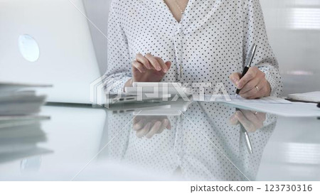 Unknown businesswoman using a calculator to crunch numbers and jotting down meticulous notes at a sleek glass desk in a contemporary office, closeup view. Taxes, audit in business 123730316