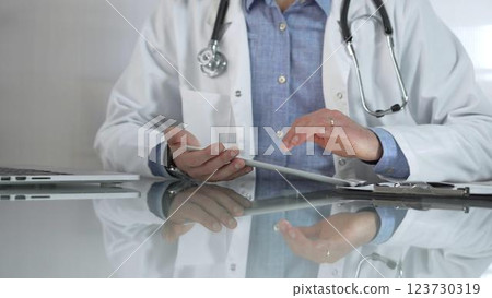 Doctor man, wearing a blue shirt under a medical white coat, is using digital tablet in modern healthcare setting, interacting with patient data, close-up. Medicine concept 123730319