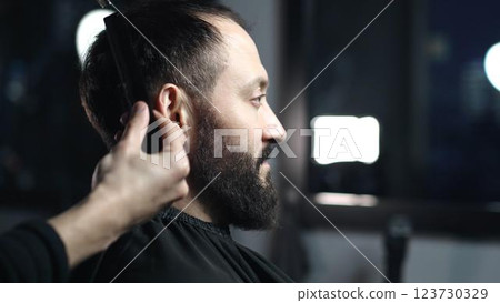 Professional barber trimming client's hair using professional scissors, creating stylish fade for bearded man in modern barbershop setting Professional barber trimming client's hair using professional scissors, creating stylish fade for bearded man in modern barbershop setting 123730329