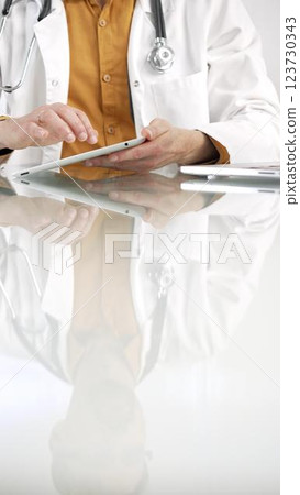 Unknown male doctor wearing a stethoscope, yellow shirt under a lab coat, is using digital tablet in modern healthcare setting, interacting with patient data, vertical portrait. Medicine concept Unknown male doctor wearing a stethoscope, yellow shirt under a lab coat, is using digital tablet in modern healthcare setting, interacting with patient data, vertical portrait. Medicine concept 123730343