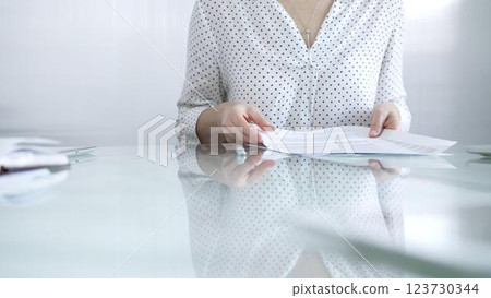 Businesswoman in white dotted blouse is going through paperwork while holding documents at a glass desk in a well lit, contemporary office setting. Business people concept Businesswoman in white dotted blouse is going through paperwork while holding documents at a glass desk in a well lit, contemporary office setting. Business people concept 123730344