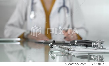 Stethoscope and medication history records form are on a glass table before a female doctor in yellow blouse under a white medical coat. Physician is working on clinical office background. Medicine Stethoscope and medication history records form are on a glass table before a female doctor in yellow blouse under a white medical coat. Physician is working on clinical office background. Medicine 123730346