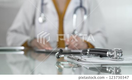 Stethoscope and medication history records form are on a glass table before a female doctor in yellow blouse under a white medical coat. Physician is working on clinical office background. Medicine Stethoscope and medication history records form are on a glass table before a female doctor in yellow blouse under a white medical coat. Physician is working on clinical office background. Medicine 123730347