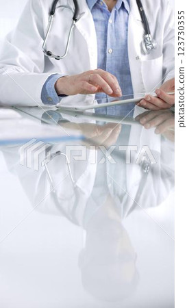 Unrecognizable doctor man in blue shirt under a white coat using digital tablet in modern healthcare setting, interacting with patient data, closeup vertical view. Medicine and health care concept Unrecognizable doctor man in blue shirt under a white coat using digital tablet in modern healthcare setting, interacting with patient data, closeup vertical view. Medicine and health care concept 123730355