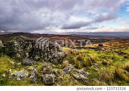 A large rock sits in the foreground, surrounded by grass and scattered stones. In the distance, rolling hills stretch out under a clear sky, showing the beauty of nature. A large rock sits in the foreground, surrounded by grass and scattered stones. In the distance, rolling hills stretch out under a clear sky, showing the beauty of nature. 123731526