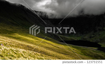 Lush green hills contrast with dark peaks shrouded in clouds, while a tranquil lake lies quietly below, embodying the wild beauty of Ireland's landscape. 123731551