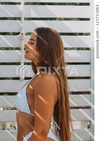 A woman with long brown hair is standing in front of a white wall. She is wearing a white bikini and she is enjoying the sun 123731663