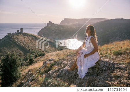 A woman in a white dress sits on a rock overlooking a body of water. The scene is serene and peaceful, with the woman enjoying the view and the calmness of the surroundings. A woman in a white dress sits on a rock overlooking a body of water. The scene is serene and peaceful, with the woman enjoying the view and the calmness of the surroundings. 123731694