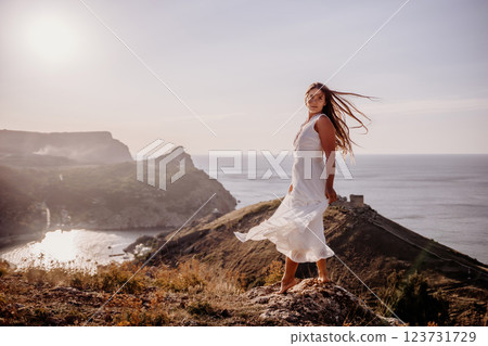 A woman in a white dress stands on a hill overlooking the ocean. The scene is serene and peaceful, with the woman's dress billowing in the wind. The combination of the ocean. 123731729