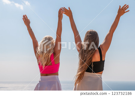Two women are standing on a beach, one wearing a pink top and the other wearing a black top. They are both holding their hands up in the air, as if they are celebrating something. The sky is clear. Two women are standing on a beach, one wearing a pink top and the other wearing a black top. They are both holding their hands up in the air, as if they are celebrating something. The sky is clear. 123732186