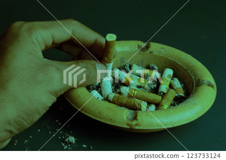 Hand extinguishing cigarette, cigarette butt in ashtray, dark green background. Hand extinguishing cigarette, cigarette butt in ashtray, dark green background. 123733124