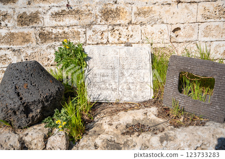 Old stone inscription with Arabic script resting against weathered wall in Northern Cyprus. Ancient artifact, surrounded by grass and volcanic stones, reflects the region historical and cultural depth Old stone inscription with Arabic script resting against weathered wall in Northern Cyprus. Ancient artifact, surrounded by grass and volcanic stones, reflects the region historical and cultural depth 123733283