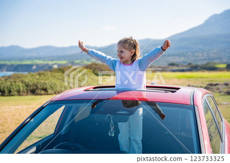 Little girl joyfully sticks out of car sunroof with raised hands against the backdrop of mountains in Northern Cyprus. The image symbolizes freedom, adventure, and the excitement of travel with child Little girl joyfully sticks out of car sunroof with raised hands against the backdrop of mountains in Northern Cyprus. The image symbolizes freedom, adventure, and the excitement of travel with child 123733325